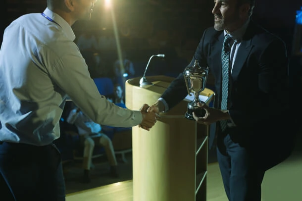 Two men shake hands on stage in Austin, celebrating a PR victory; one holds a trophy, symbolizing success in PR management and consulting.